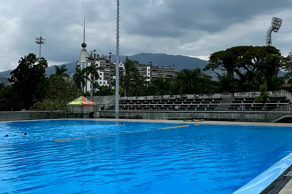 La piscina olímpica del Atanasio Girardot, azul intenso, con las graderías, las palmas y la montaña de Medellín al fondo bajo un cielo nublado.