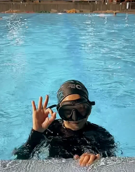 Una makara con gorro negro del club y máscara, apoyada en el borde de la piscina, hace la señal de OK